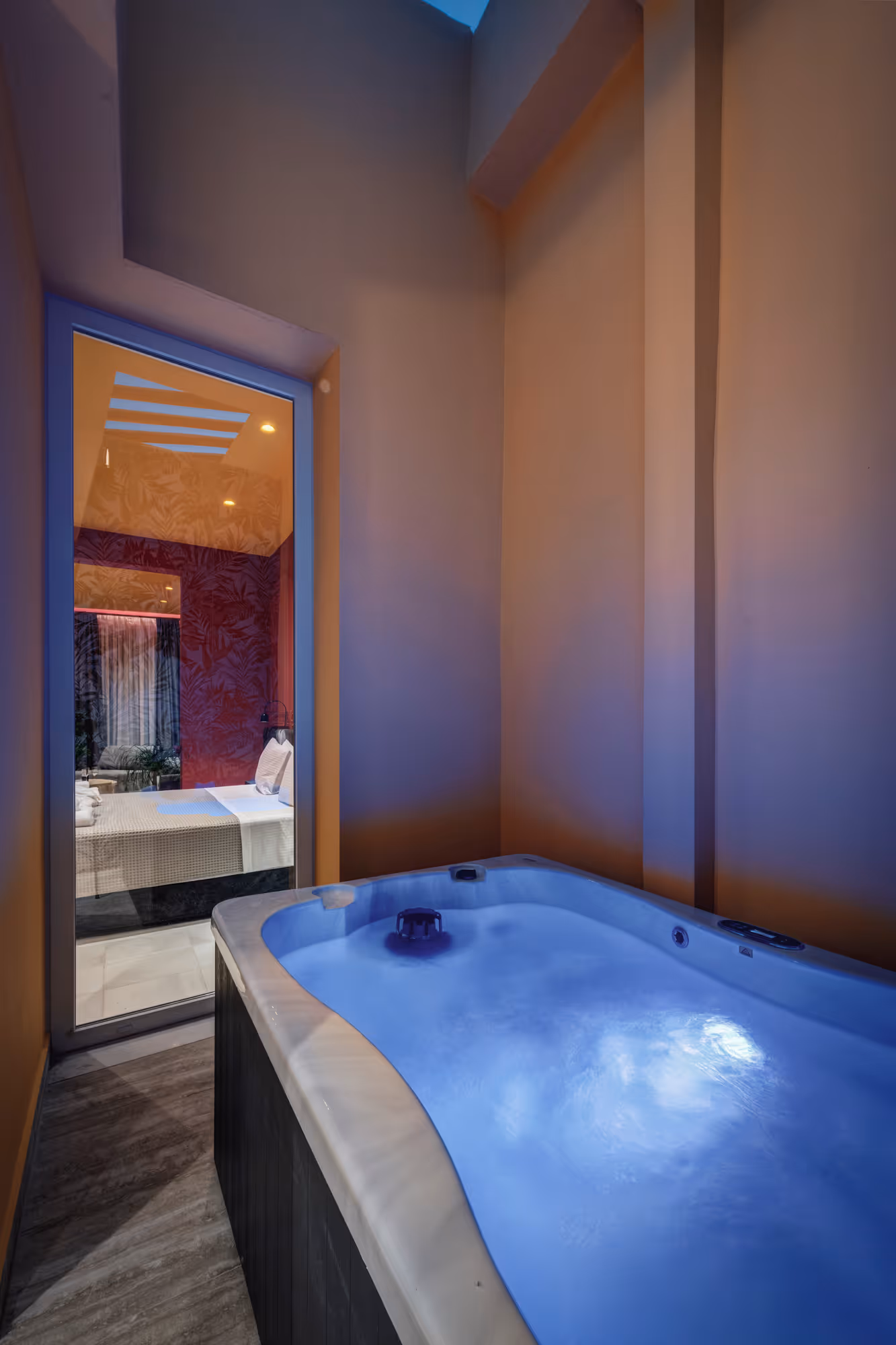 Outdoor jacuzzi with blue illuminated water beside a glass door showing a bedroom with a bed and textured wallpaper.