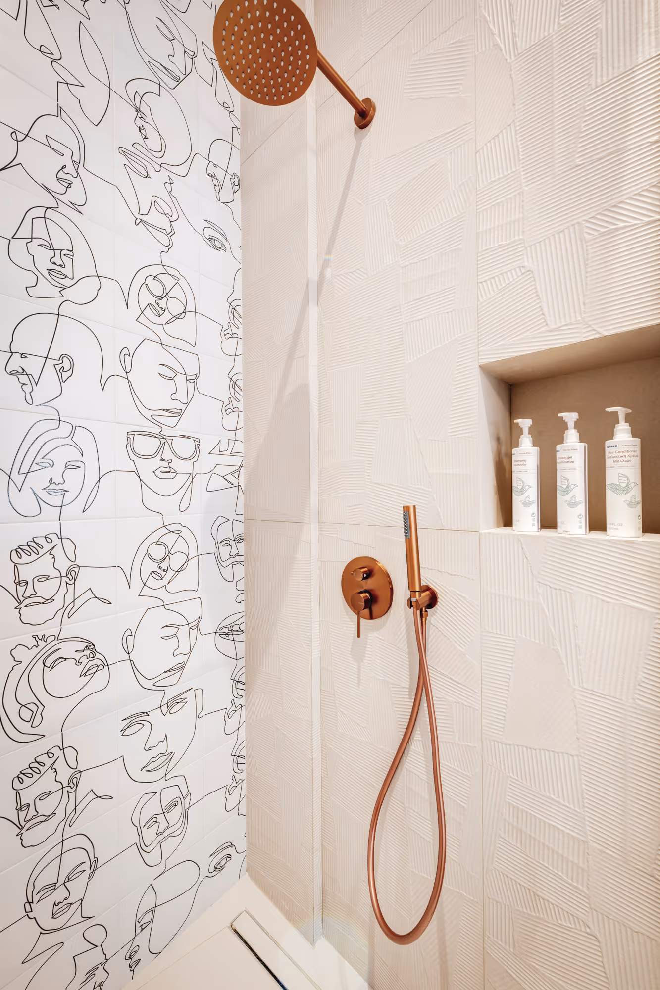 Modern shower with copper fixtures, textured beige tiles, and white wall tiles featuring black continuous line art faces, and three white pump bottles in a built-in niche.