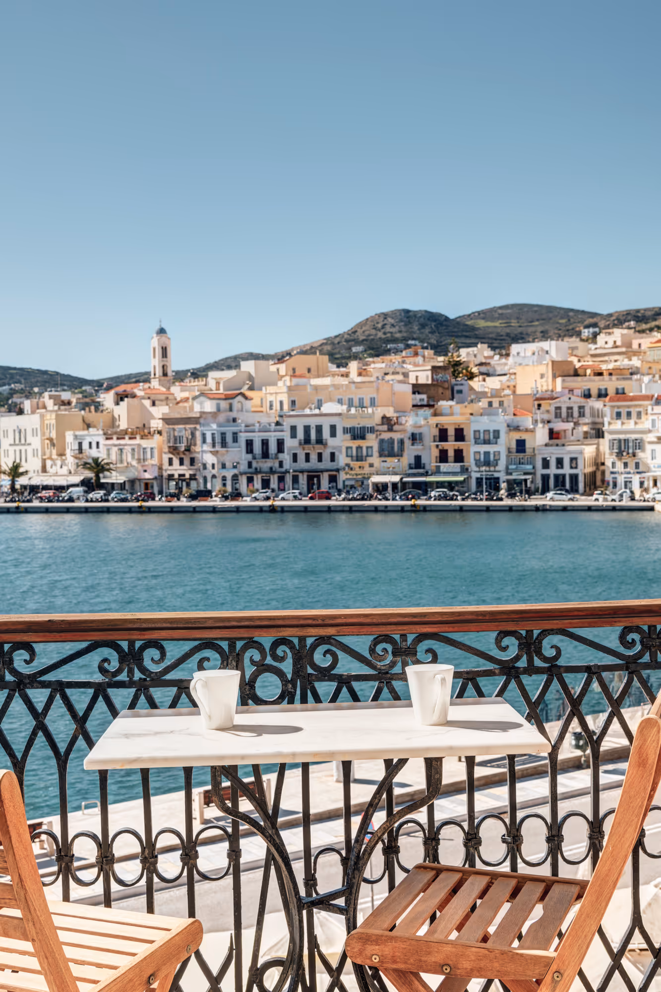 Two wooden chairs and a small table with two white mugs on a balcony overlooking a calm blue waterfront and a town with light-colored buildings and hills in the background.