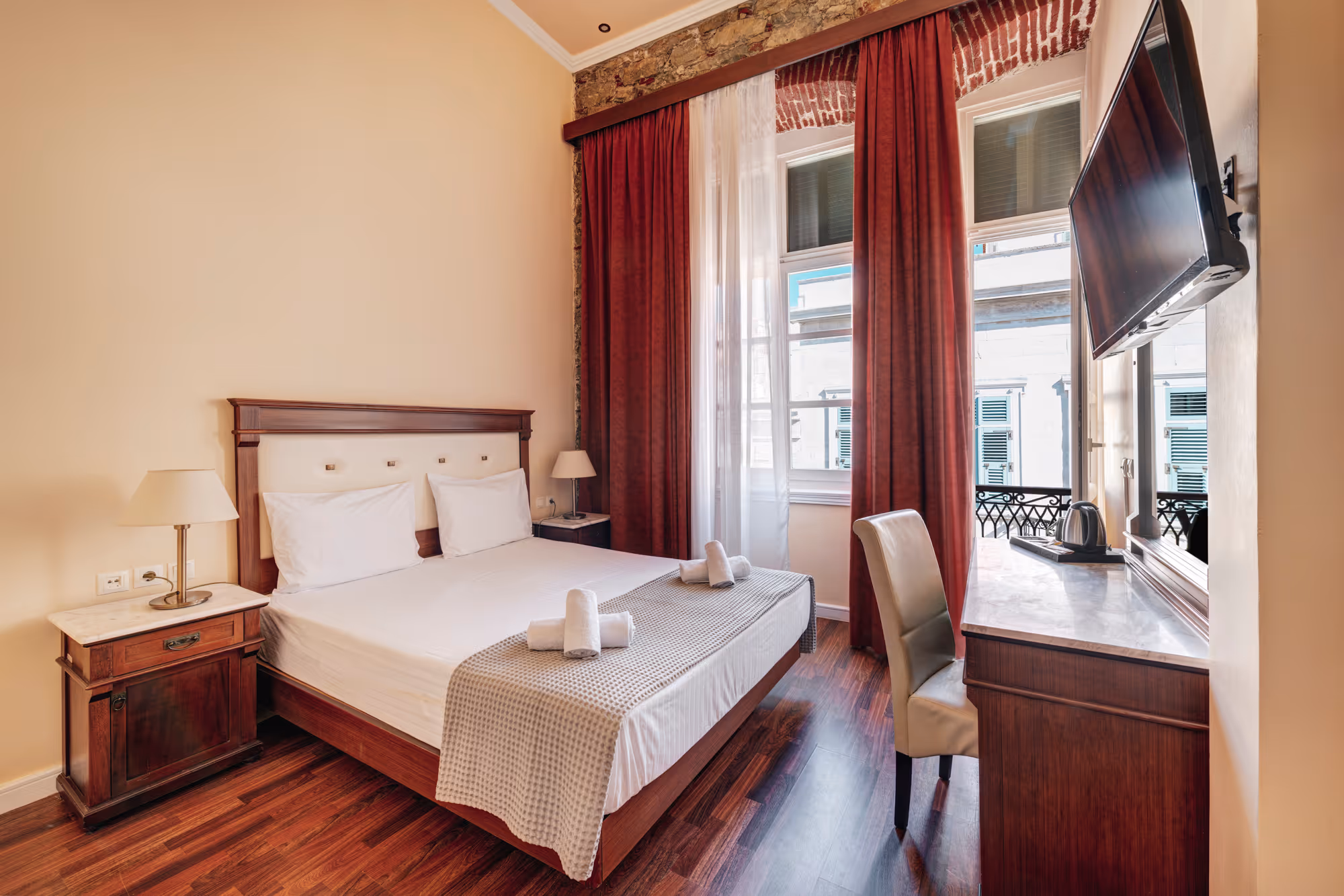 Hotel room with wooden double bed, white linens, rolled towels, red curtains, a desk with a chair, and a wall-mounted TV.
