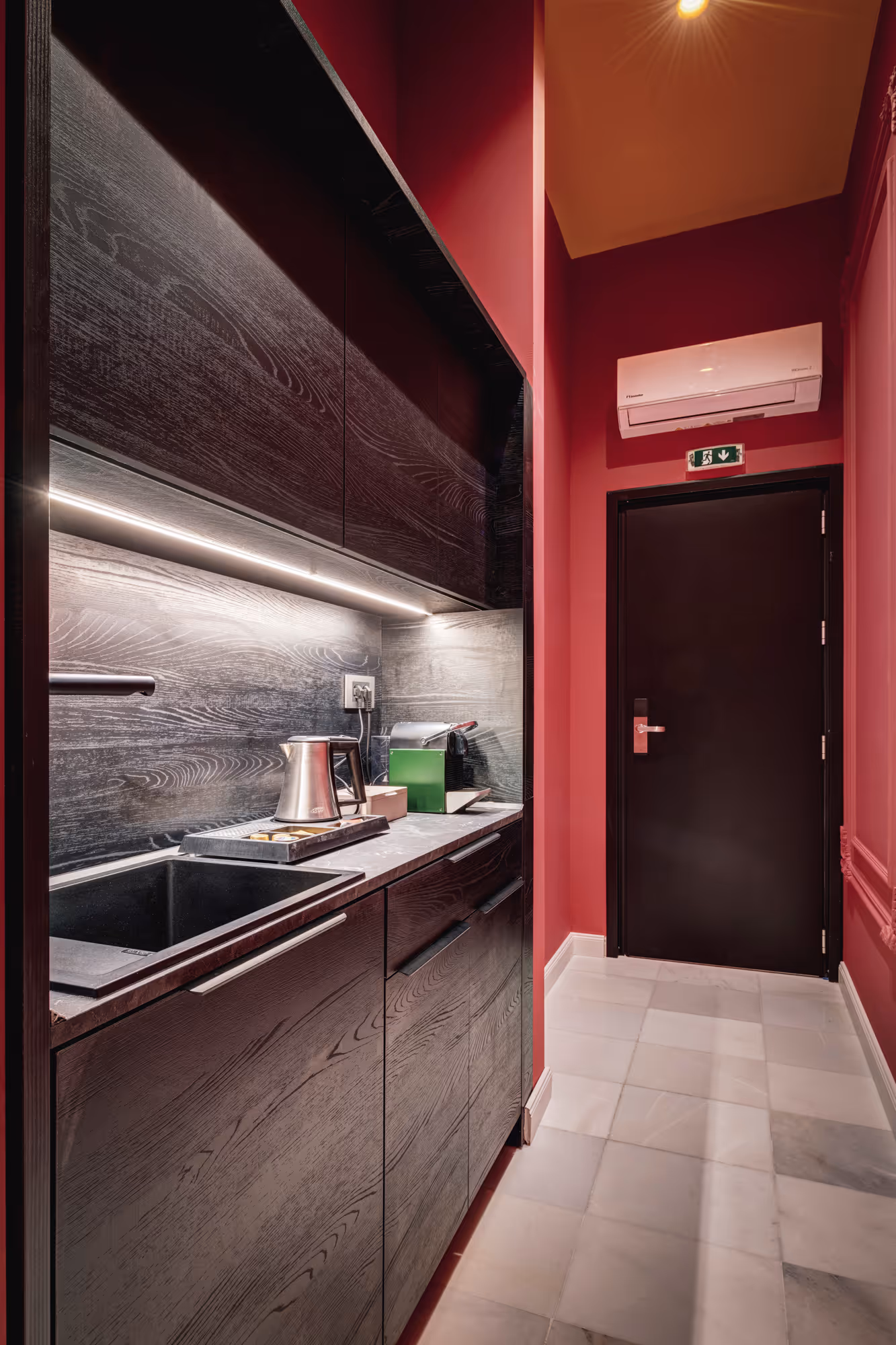 Modern kitchenette with dark wood cabinets, under-cabinet lighting, a sink, electric kettle, coffee maker, and a black door at the corridor end with red walls and white tiled floor.