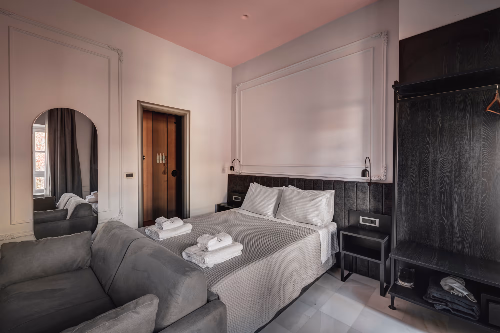 Modern hotel room with a double bed, two folded towels on the bed, gray sofa, full-length mirror, and dark wood open wardrobe.