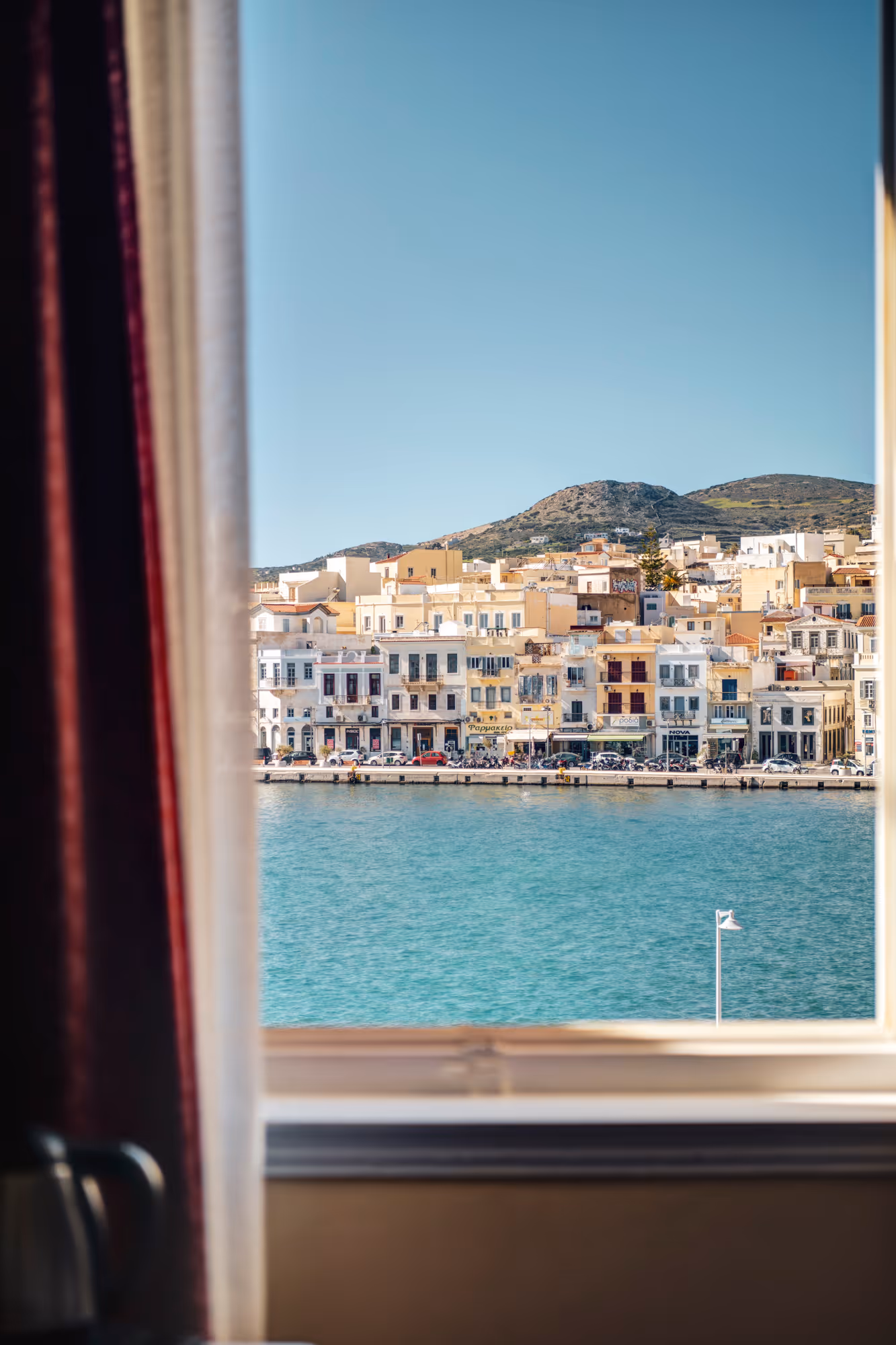 View of a seaside town with pastel-colored buildings, blue water, and hills in the background, seen through a window with curtains.