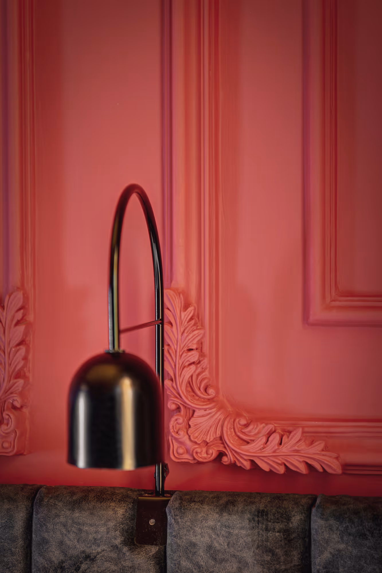 Black wall-mounted curved lamp above a dark textured couch against a detailed pink wall with ornate molding.