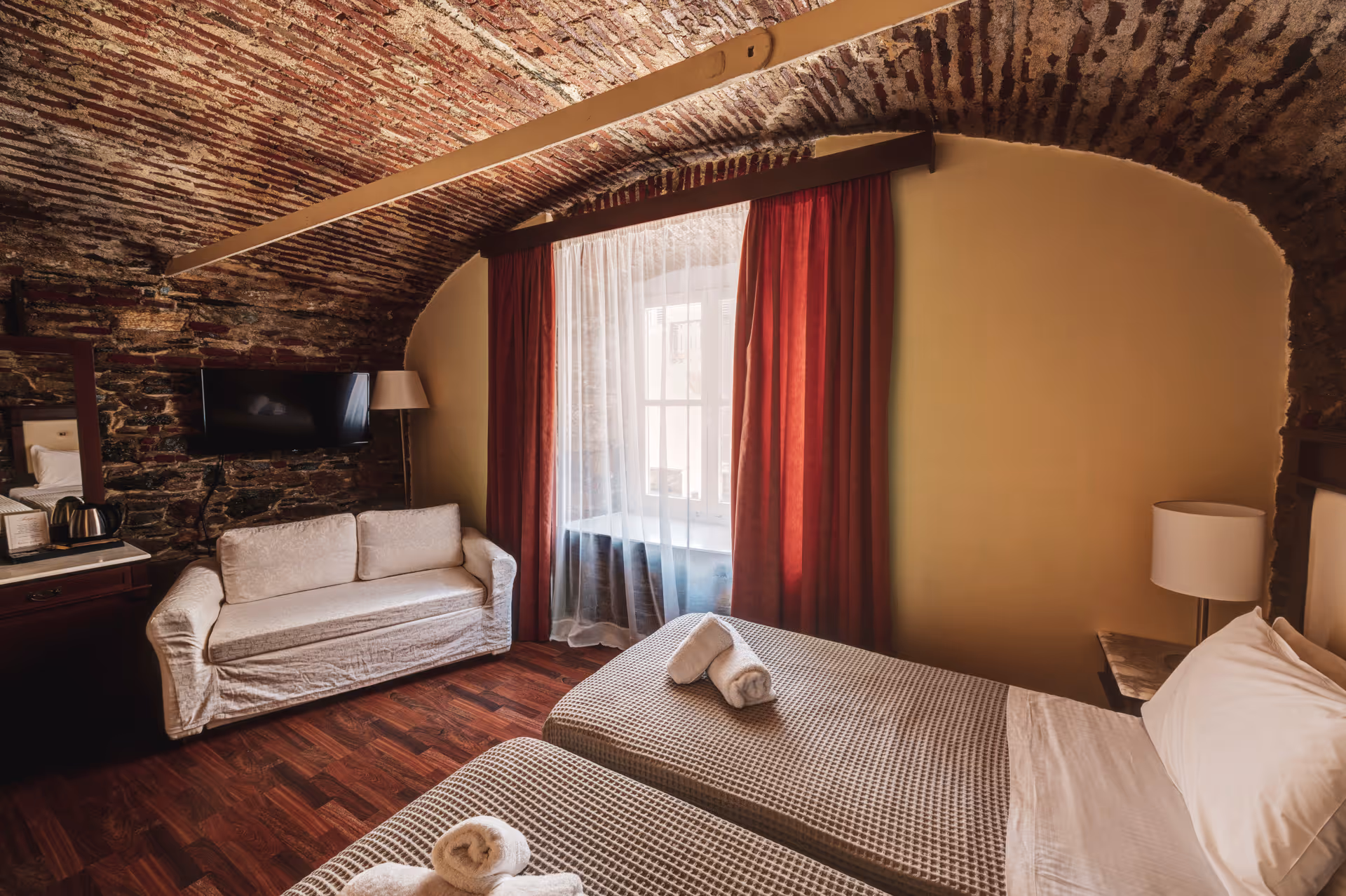 Cozy hotel room with two single beds, rolled white towels, a white sofa, brick arched ceiling, and a window with red curtains.