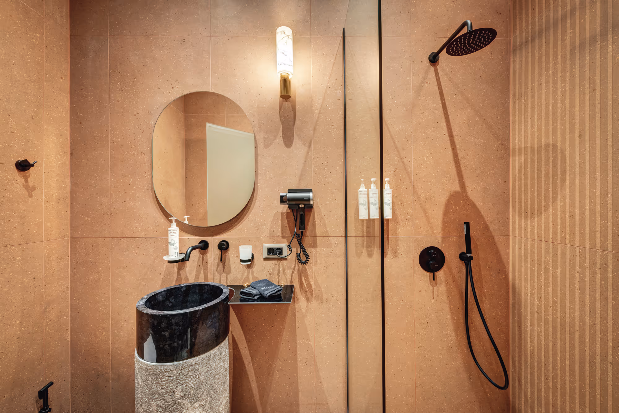 Modern bathroom with pinkish beige tiled walls, oval mirror, black freestanding sink, wall-mounted faucet, hairdryer, and black rain showerhead with handheld sprayer.