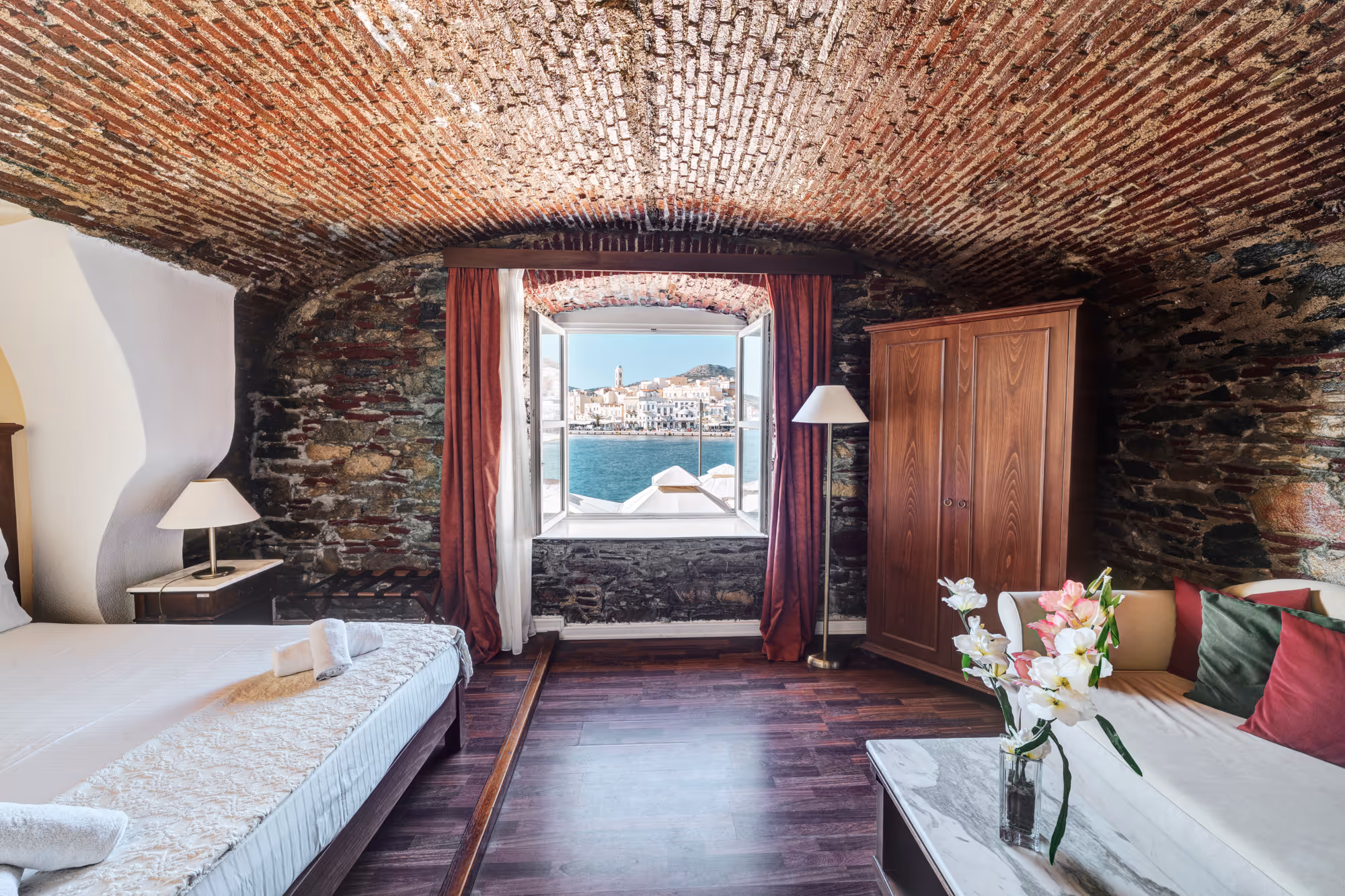 Cozy room with arched brick ceiling, bed with white linens, wooden wardrobe, sofa, and open window overlooking sea and town.