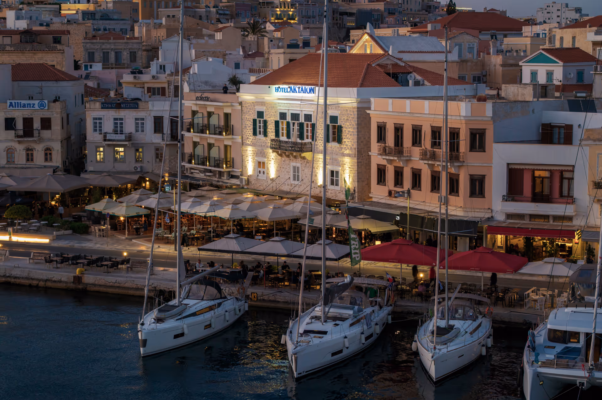Harbor at dusk with sailboats docked in front of a waterfront lined with illuminated outdoor cafes and historic buildings.