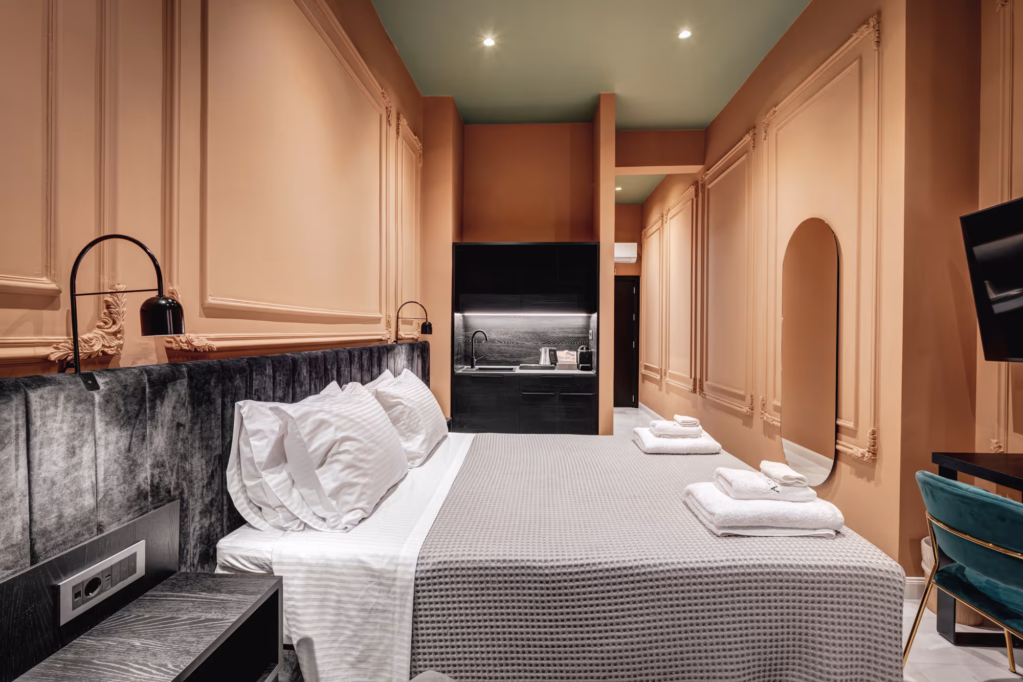 Modern hotel room with a large bed, gray textured headboard, soft peach walls with decorative molding, and a small kitchenette in the background.