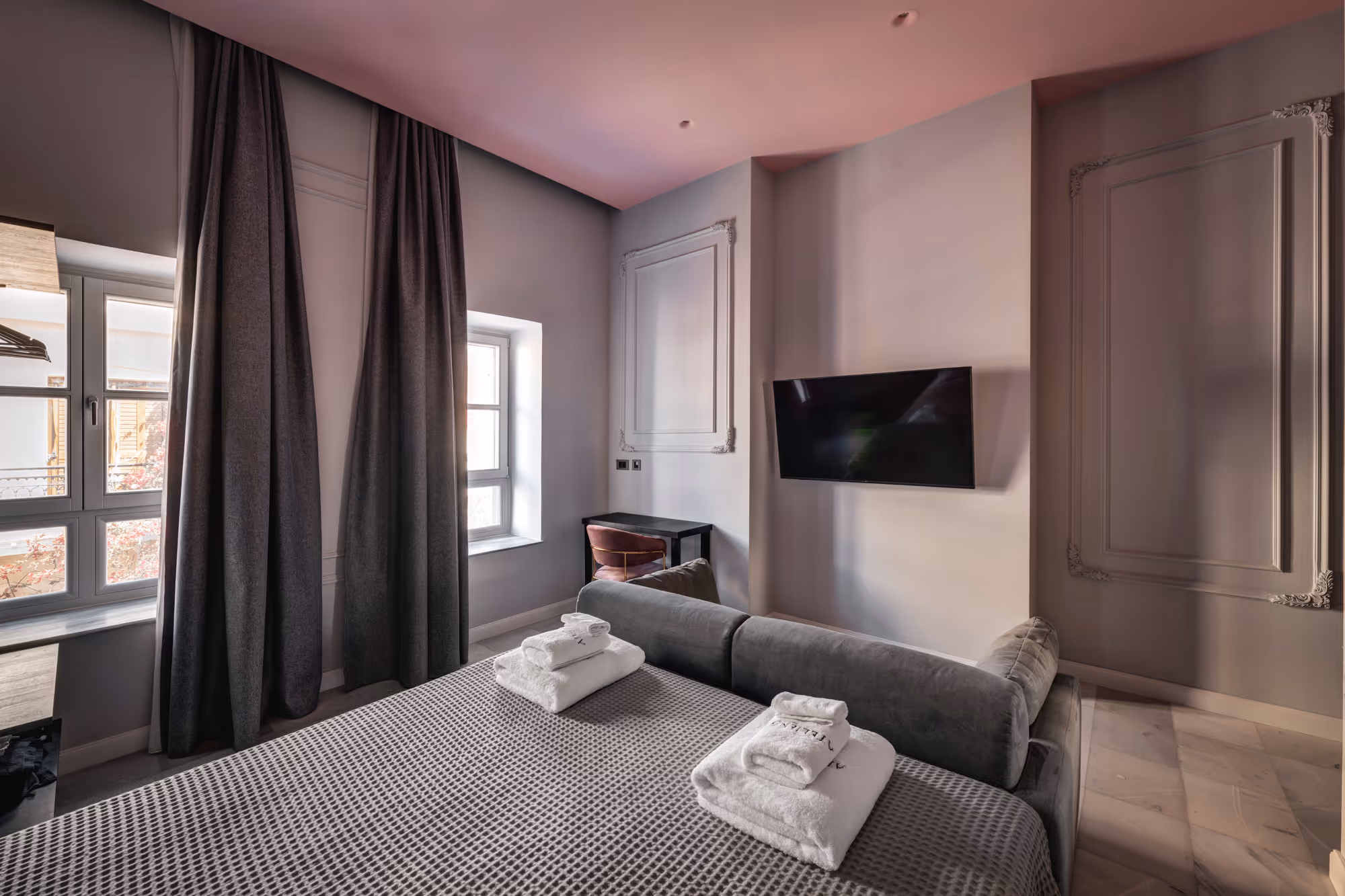 Modern hotel room with a gray bed, folded white towels, large windows with dark curtains, a mounted flat-screen TV, and a small desk with a chair.